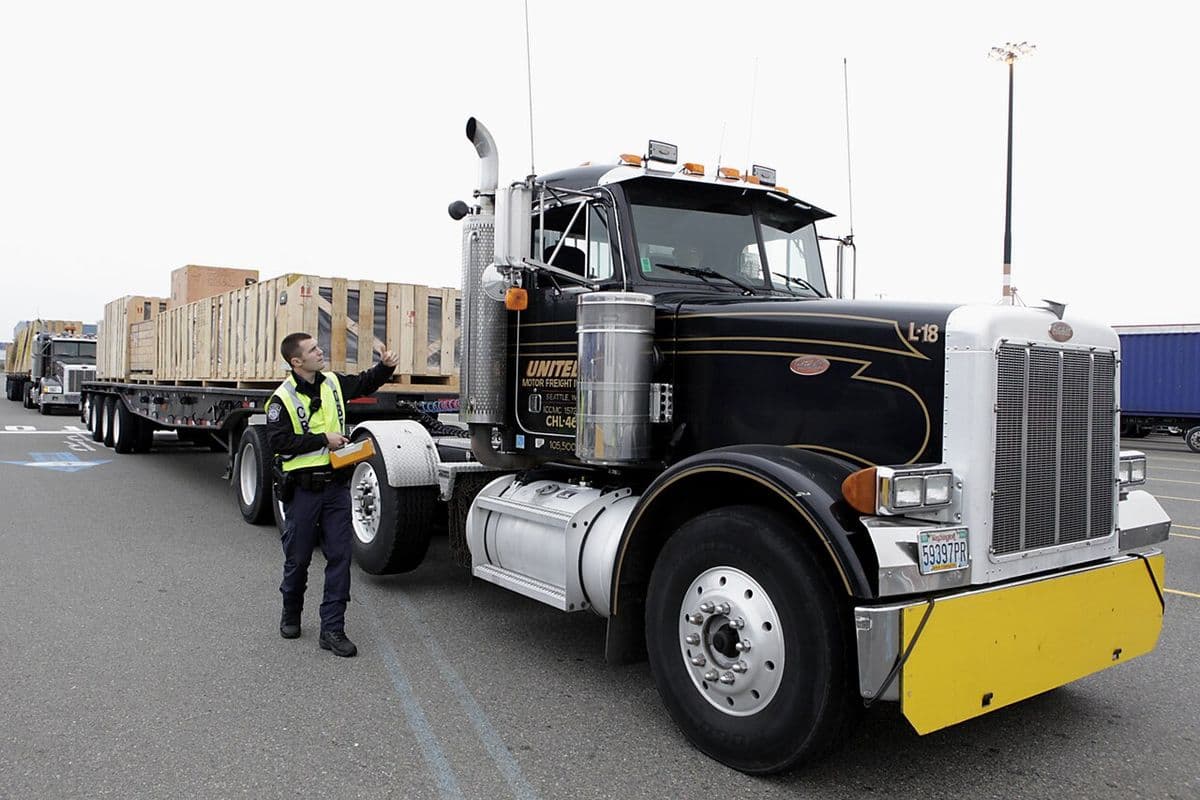 An officer in a safety vest signals a flatbed truck loaded with wooden crates through an inspection station in Seattle, Washington.