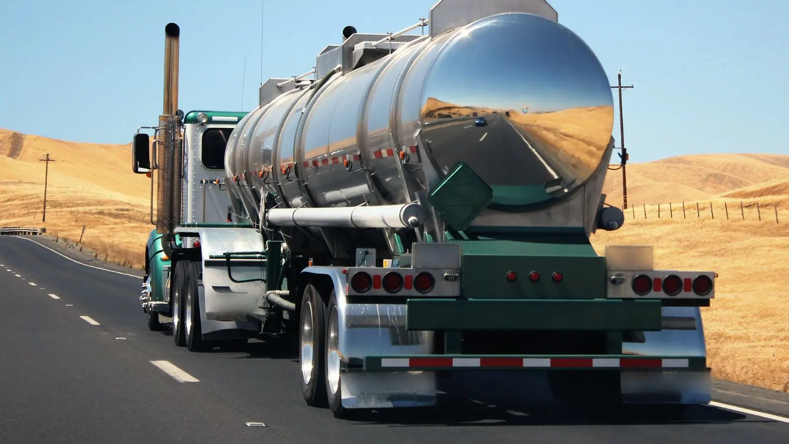 A polished tanker truck drives along a divided highway through dry golden hills in California.