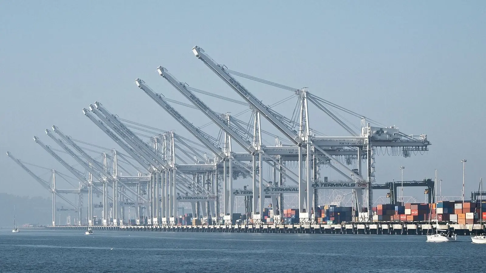 A long row of container cranes lines the waterfront at the Port of Oakland in California, with stacked containers below.
