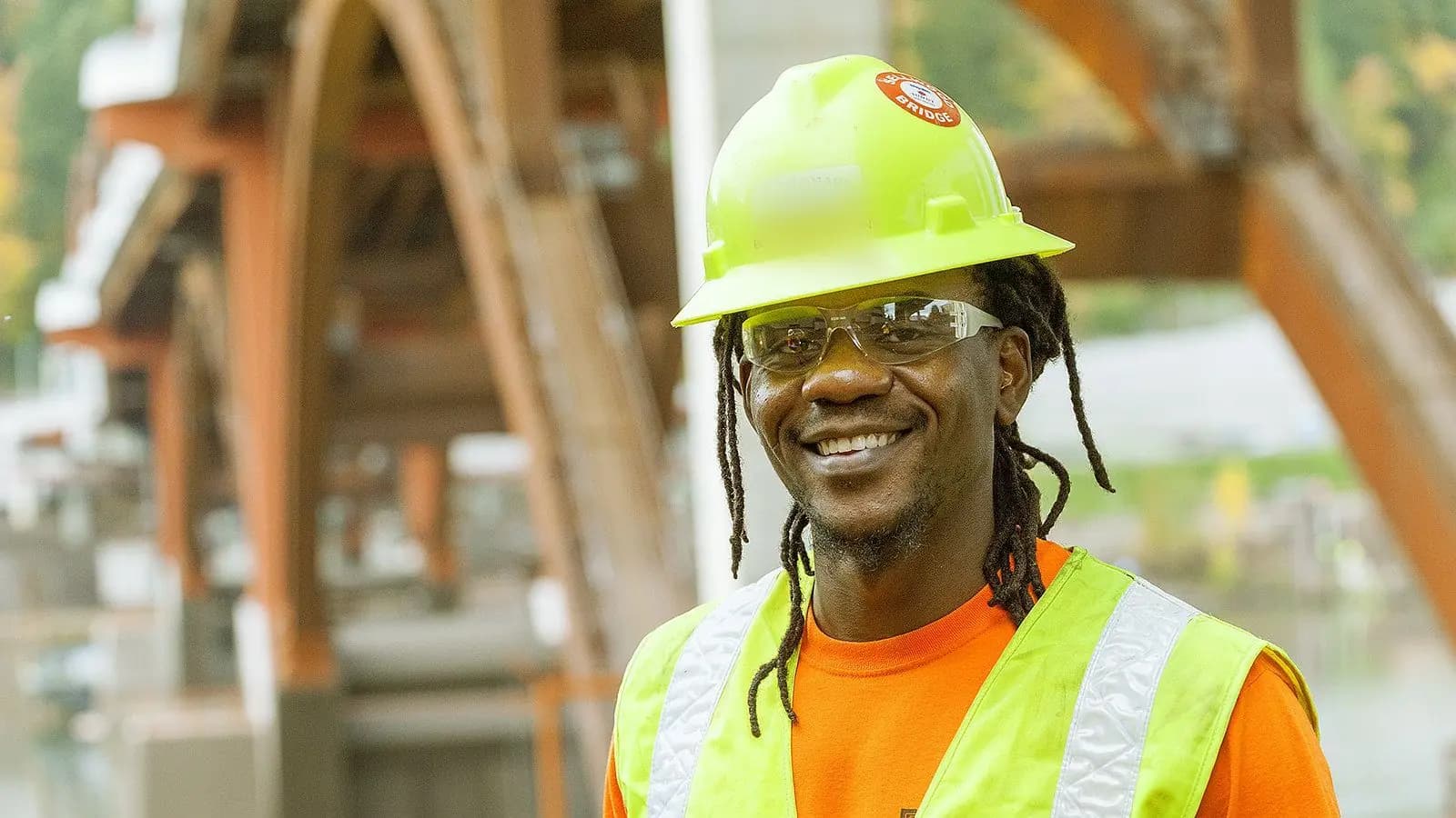 A smiling carpenter apprentice in a hard hat, safety glasses and a high-visibility vest stands in front of a bridge under construction.