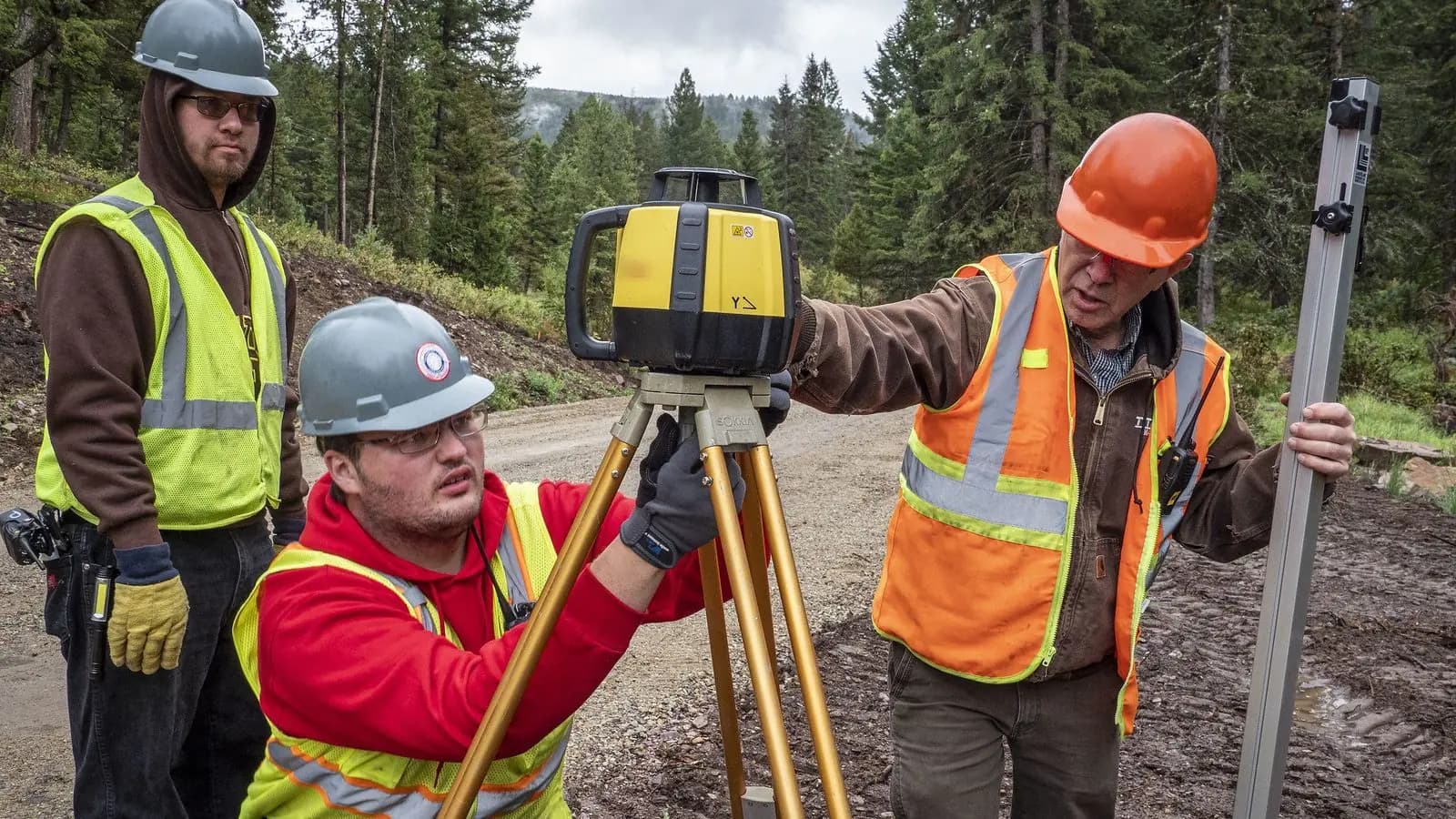 An instructor shows two trainees how to set up a laser level on a tripod beside a gravel forest road in Montana.