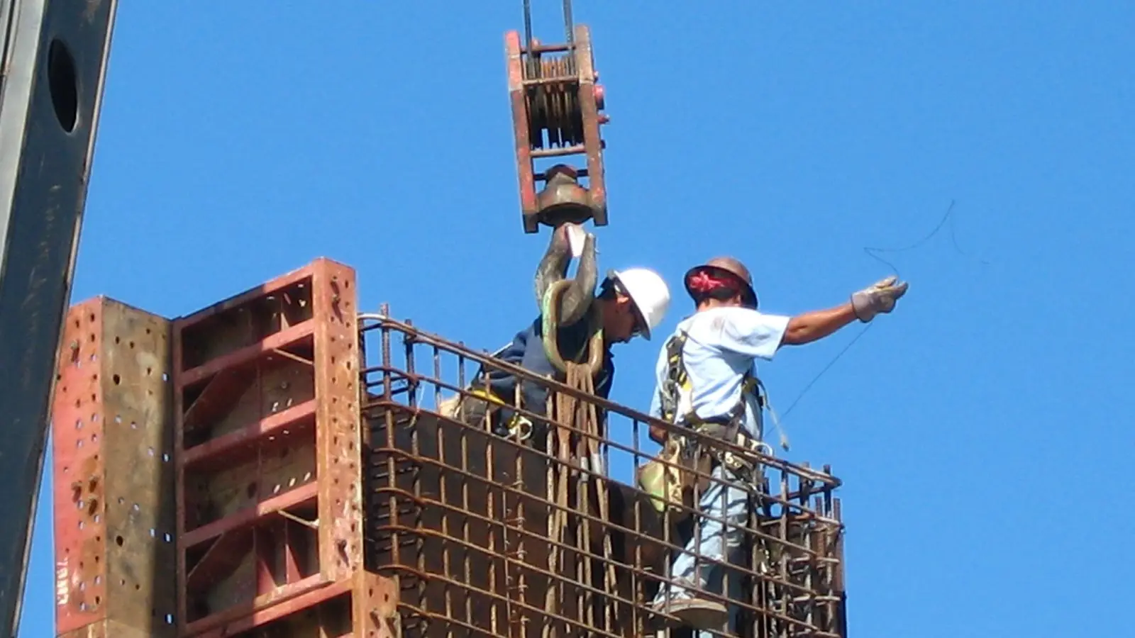 Two construction workers on top of a steel rebar cage guide a crane hook against a clear blue sky at a Houston, Texas, freeway project.